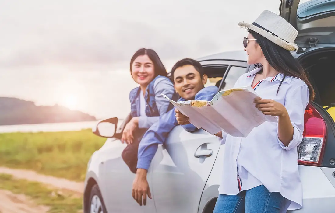Happy-asian-woman-and-her-friends-standing-by-car-on-coastal-road-at-sunset for outdoor tour
